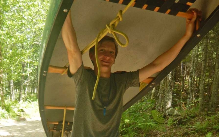 A person carries an upside-down canoe on their shoulders through a green wooded area